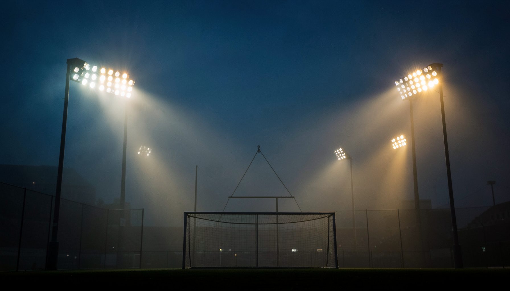 Stadium floodlights piercing through evening mist, dramatic silhouette of goalposts, deep blue and gold hues
