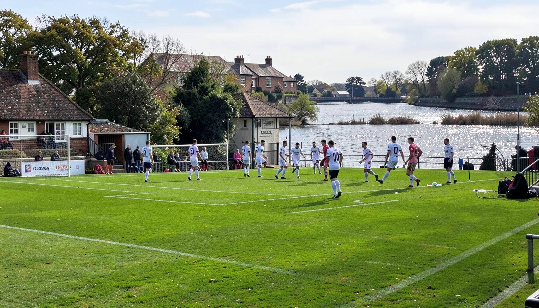 Craven Cottage seen from the Riverside Stand, the Thames glinting behind, Fulham players in white shirts warming up on the pitch before kick-off