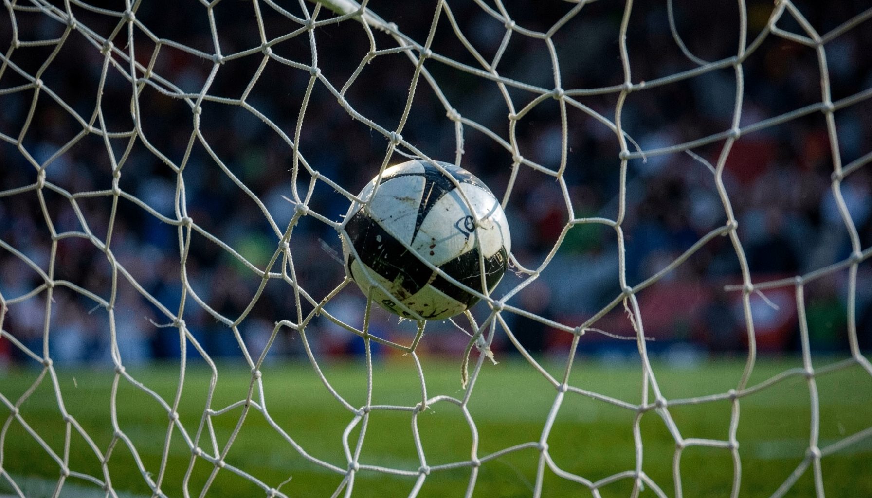 Football net rippling as a ball strikes it, stadium seats in soft focus behind, dramatic evening lighting