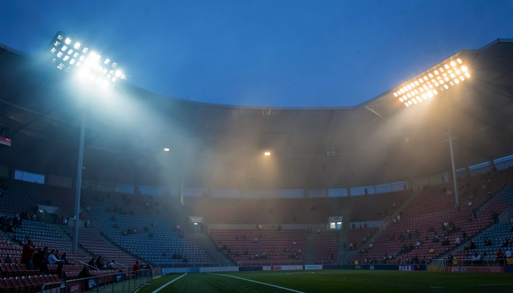 Stadium floodlights piercing through evening mist, cool blue and warm amber light blending across the stands
