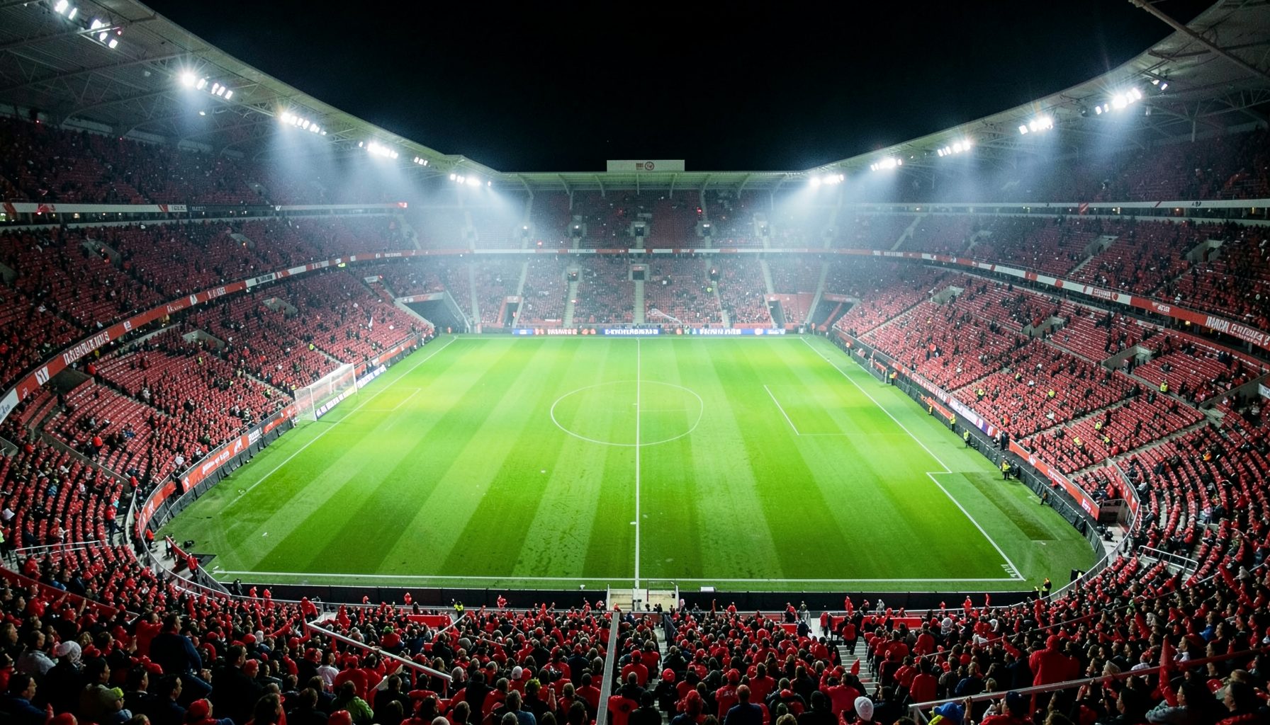 A wide aerial view of a packed English football stadium under floodlights at night, the pitch a vivid green rectangle surrounded by tiers of red and white seats, with a faint mist hanging above the turf
