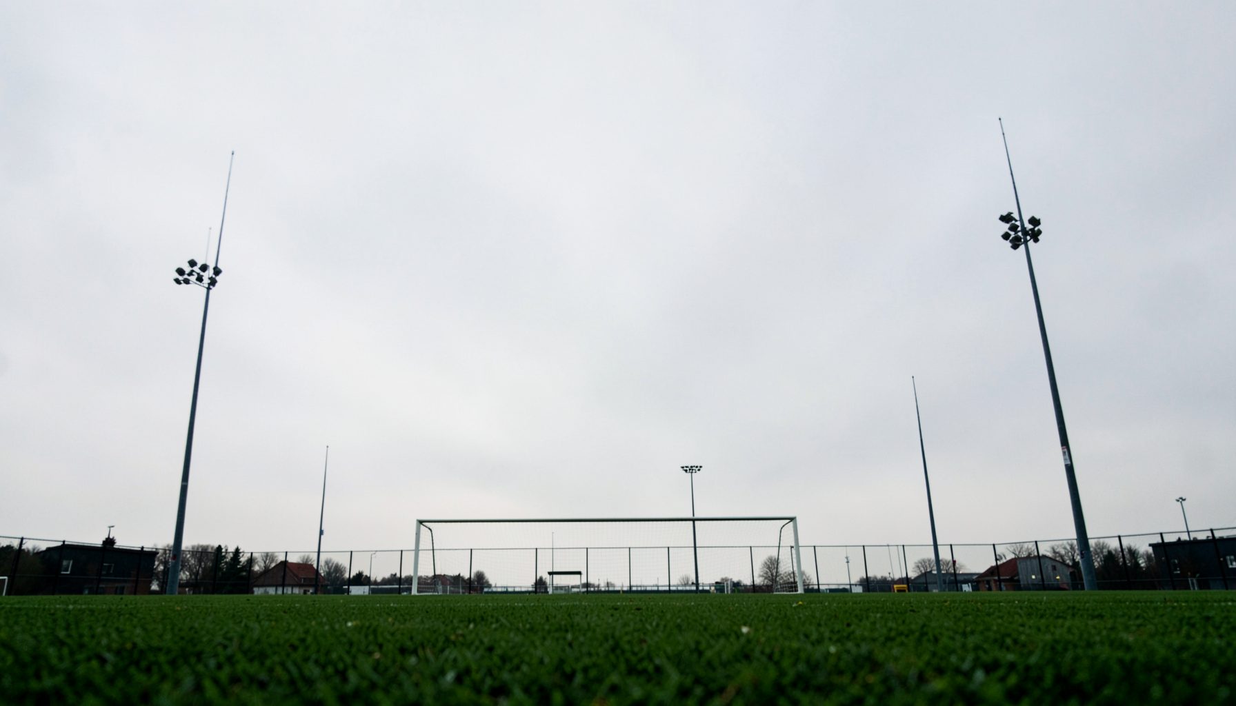 A green pitch seen from a low angle, goalposts silhouetted against a pale grey overcast sky