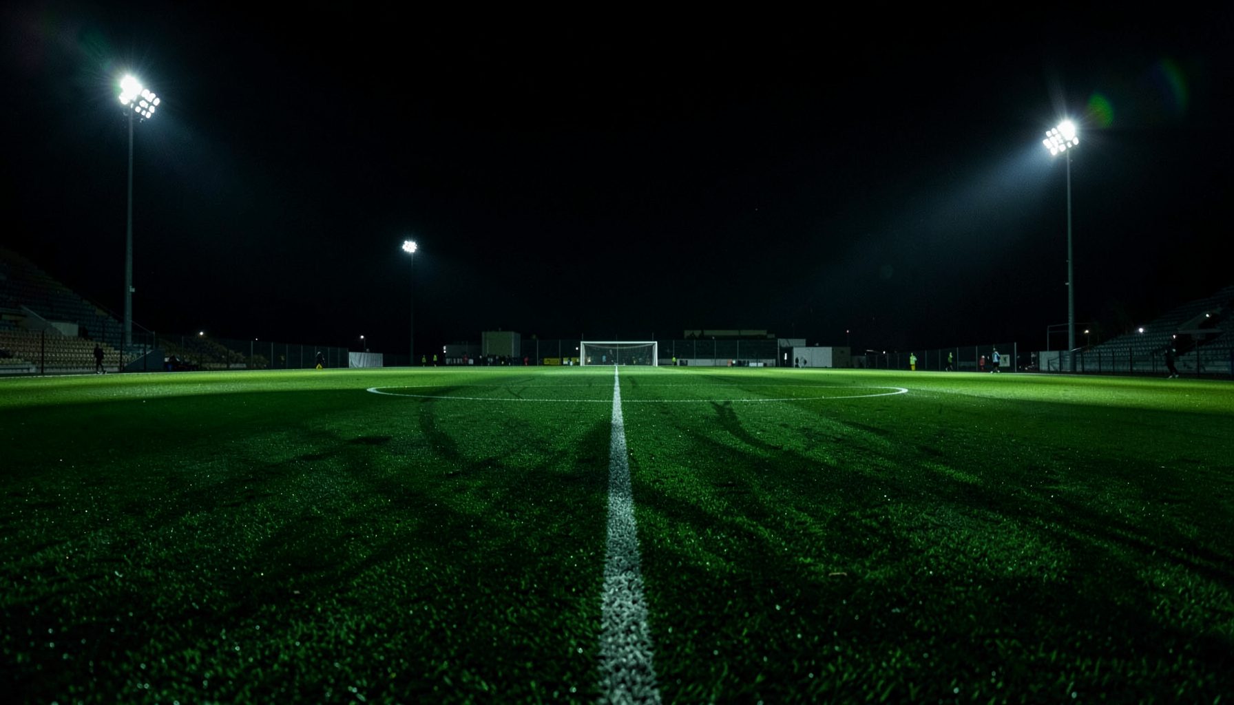Dark green football pitch at night under floodlights, dramatic shadows across the grass, intense sporting atmosphere