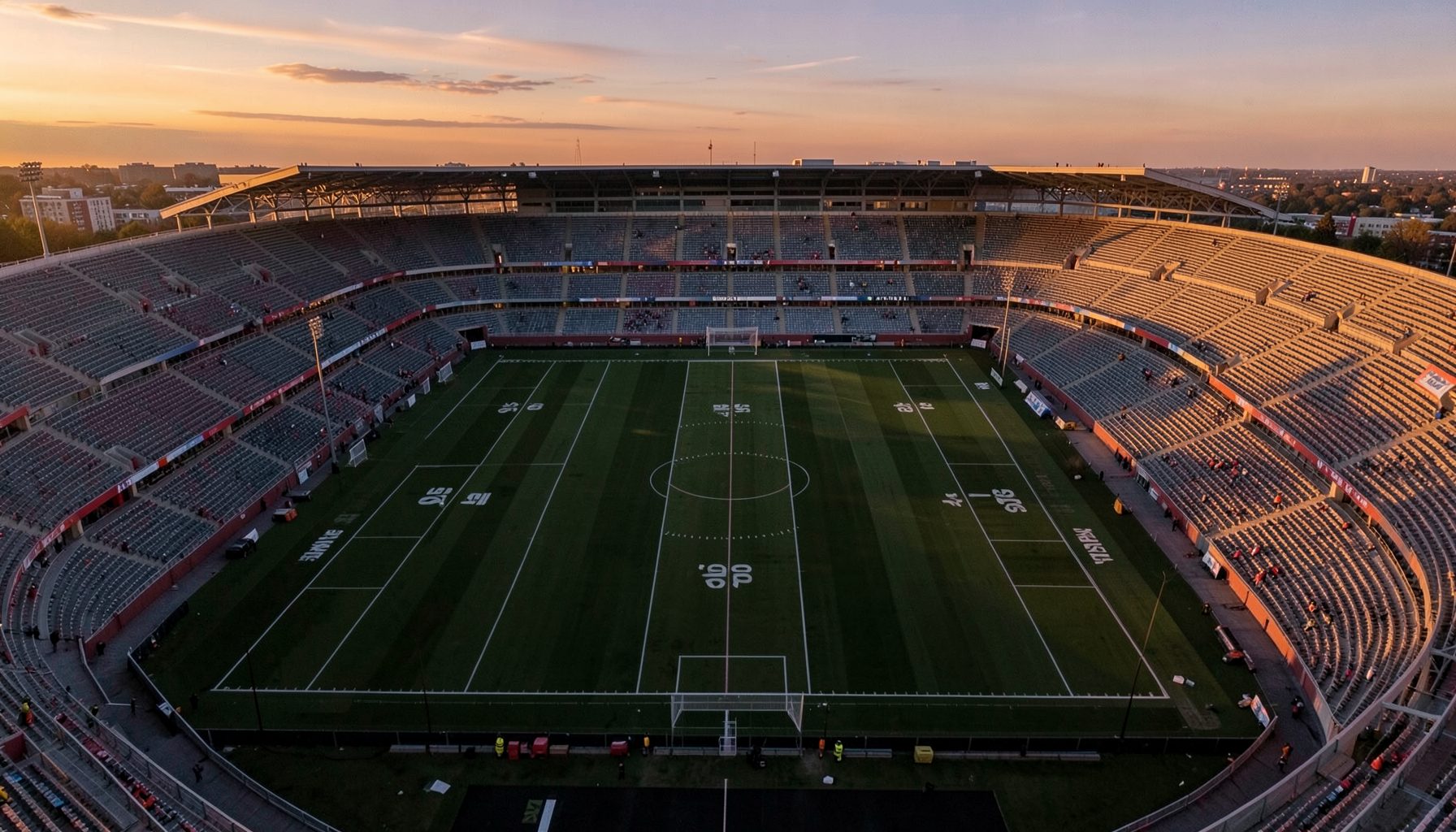 Aerial view of a football stadium at dusk, empty pitch with geometric mowing patterns, warm golden-hour light
