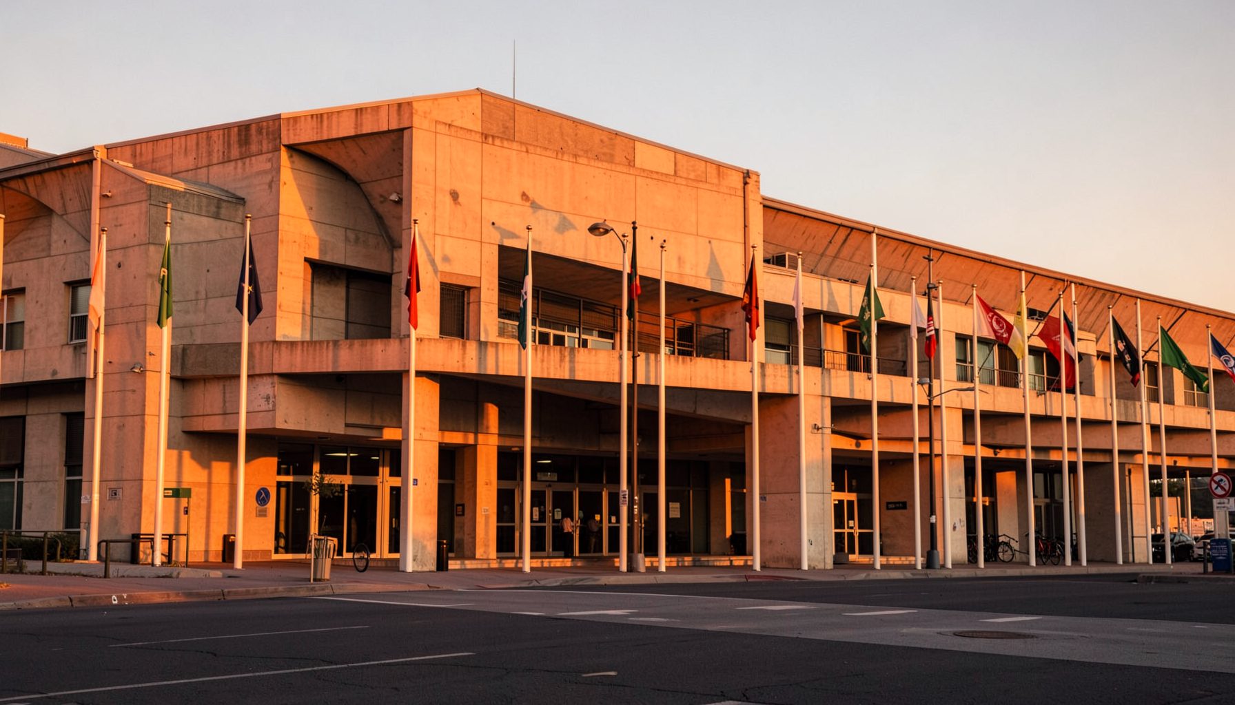 Stadium exterior at golden hour, warm orange light washing over the concrete facade, flags lining the entrance