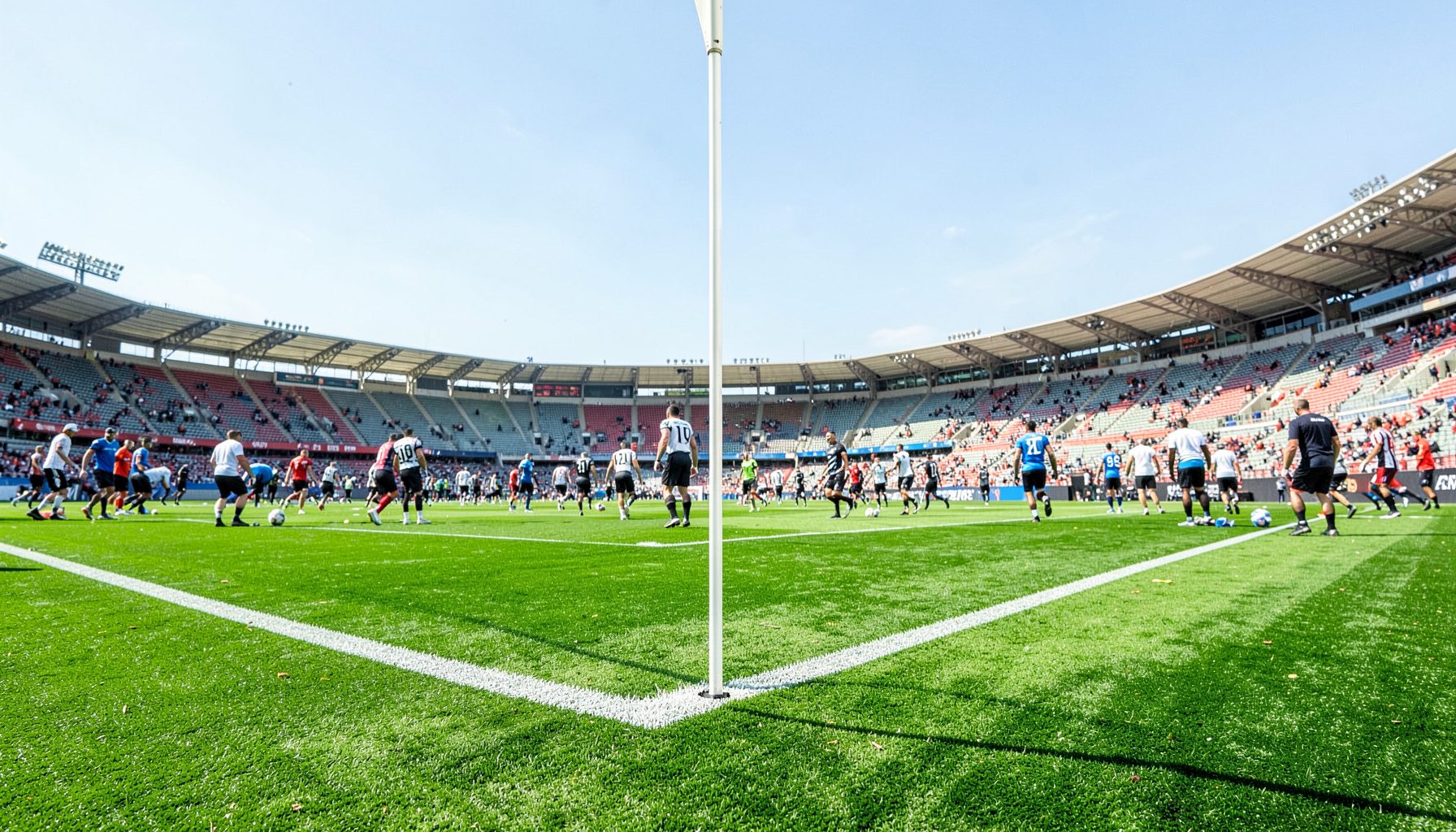 Wide shot of a football stadium corner flag with players warming up, bright afternoon light, vivid green turf