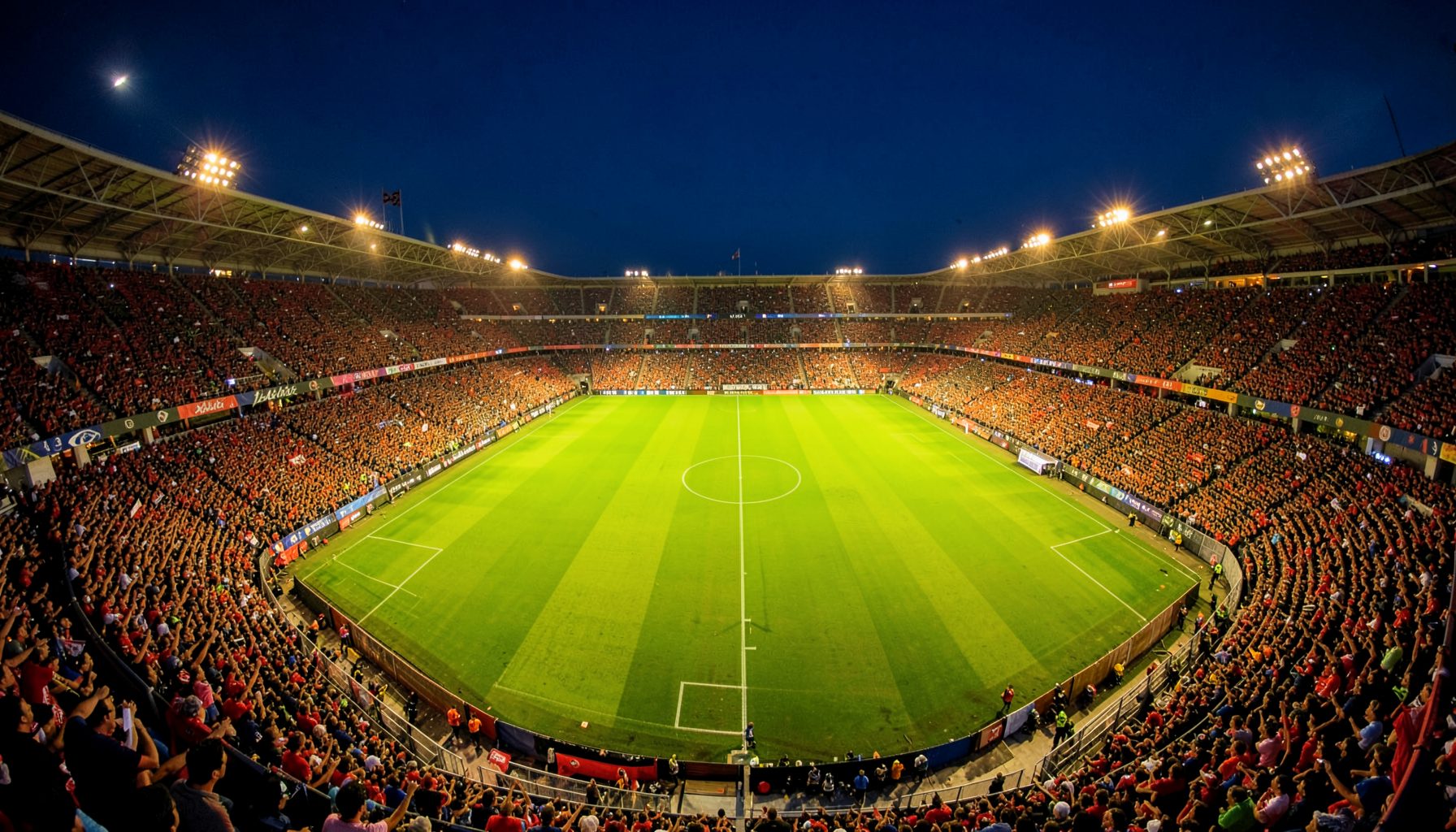 Wide aerial view of a packed Spanish football stadium at dusk, warm golden floodlights illuminating the vivid green pitch, deep navy sky above, passionate atmosphere
